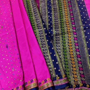 Puede incluir: Una tela rosa vibrante con intrincados bordados y adornos. El diseño presenta una combinación de acentos azul marino, verdes, dorados y rosas. La tela probablemente se utiliza para prendas tradicionales o con fines decorativos.