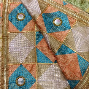Puede incluir: Primer plano de un textil colorido con un patrón geométrico. El diseño presenta cuadrados y diamantes en tonos de azul, naranja y dorado, con adornos de espejo. Los bordes están adornados con bordados dorados.