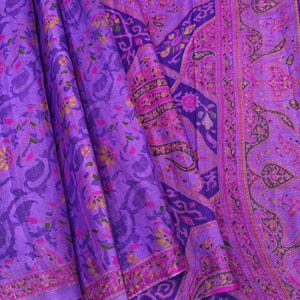 Könnte beinhalten: Ein violetter Stoff mit einem dichten Muster aus dunkelblauen Paisley-Formen und kleinen Blumenmotiven in Gelb, Pink und Grün. Das Textil zeigt einen ausgeprägten Rand mit komplizierten, sich wiederholenden Paisley-Mustern in Magenta- und Violetttönen.
