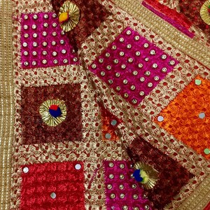 May include: Close-up of a vibrant textile with a patchwork design. The fabric features squares in shades of red, pink, orange, and brown, embellished with gold embroidery, small mirrors, and decorative accents. The edges are trimmed with a gold border.