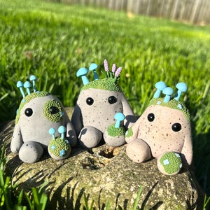 May include: Three whimsical, speckled clay figurines with mushroom and floral accents. Each has large black eyes and small arms and legs. The figurines are in shades of gray, tan, and green, sitting on a stone in a grassy area.