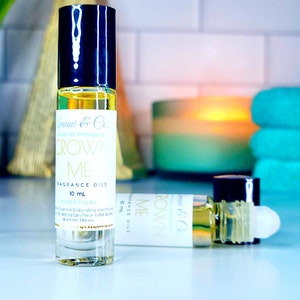 May include: A clear glass roll-on bottle with a black cap and a white label that reads "Crown Me Fragrance Oils 10 mL Directions: Apply Essential Oil by rolling directly behind Ears/Neck, Collar Bones and Inner Elbows."