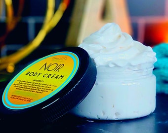 May include: A close-up of a jar of Noir Body Cream with a yellow and blue label. The cream is white and fluffy, and the jar is partially open.