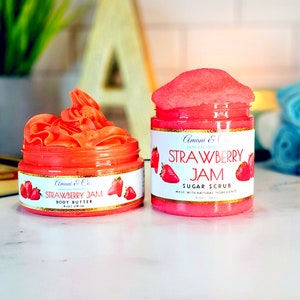 May include: Two jars of strawberry jam-themed skincare products. The jar on the left is a body butter and the jar on the right is a sugar scrub. Both jars have a gold lid and a pink and white label with the text "Strawberry Jam" and "Amani & Co."