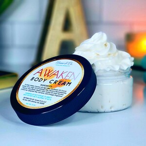 Op de afbeelding: Een zwarte pot met een witte deksel die een witte bodycrème bevat. De pot heeft een gouden etiket met de tekst "Awaken Body Cream" en een lijst met ingrediënten. De crème is geklopt en luchtig.
