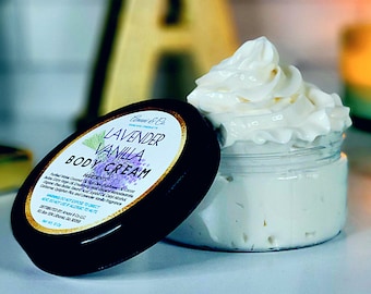 May include: A black jar with a white lid containing a white body cream. The lid has a label that reads "Lavender Vanilla Body Cream" with a purple flower graphic.