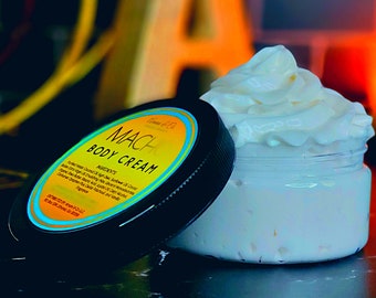 May include: A black jar of body cream with a yellow and blue label that reads "MACHI BODY CREAM". The jar is open and a dollop of white cream is on top of the jar.