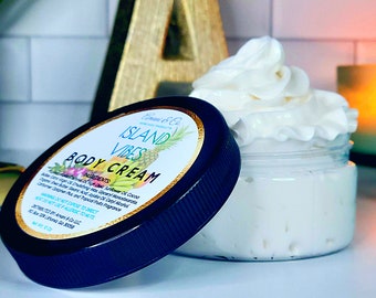 May include: A black jar with a white lid and a label that reads "Island Vibes Body Cream". The jar is filled with a white, whipped cream. The label has a pineapple and a coconut on it.