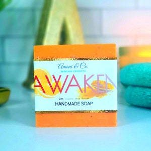 Op de afbeelding: Oranje handgemaakte zeep met een goudkleurig etiket. Het etiket leest "Amani & Co. Skincare Products" en "AWAKEN" met een sinaasappelschijfje. De zeep is gemaakt met biologische sheaboter.