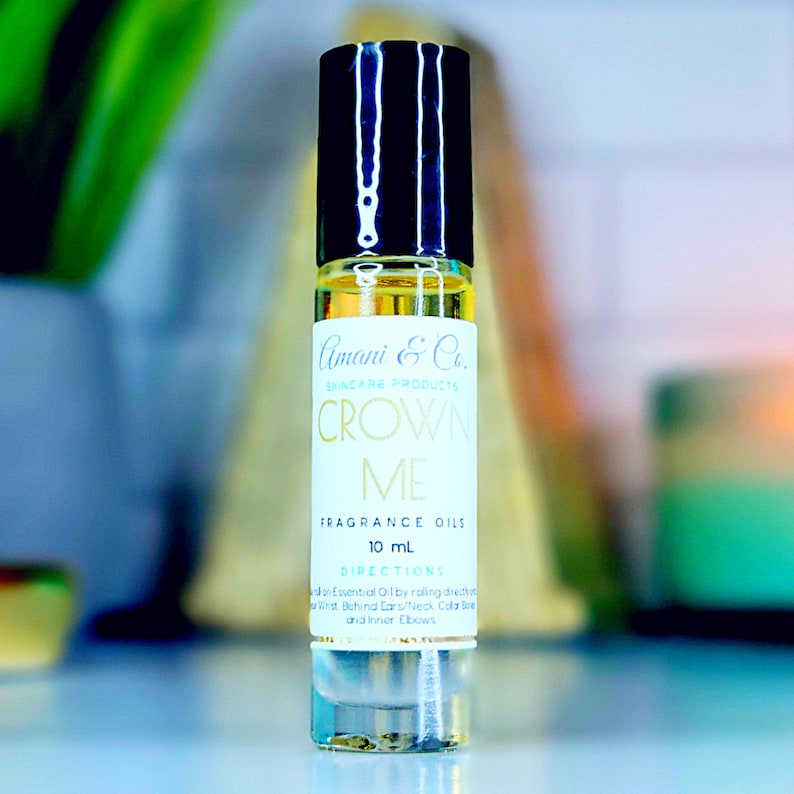 May include: A clear glass bottle with a black roll-on top. The label is white with gold text that reads "Amani & Co. Skincare Products Crown Me Fragrance Oils 10 mL Directions Apply Essential Oil by rolling directly behind Ears/Neck Collar Bone and Inner Elbows."