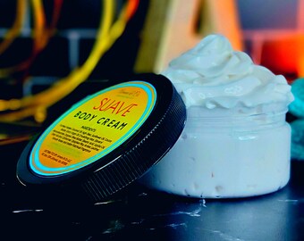 May include: A jar of Suave Body Cream with a black lid and yellow label. The label includes the product name and ingredients. The jar is filled with white, whipped cream-like body cream. The background is blurred, with hints of wood and other objects.