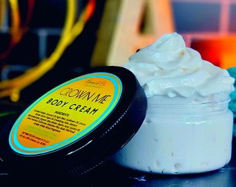 May include: A black jar with a white lid containing a white body cream. The jar has a yellow and blue label that reads "Crown Me Body Cream". The ingredients are listed on the label.