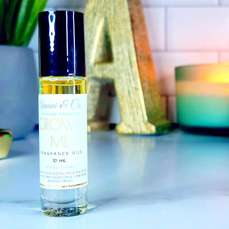 May include: A clear glass bottle with a black roll-on top. The bottle contains a yellow liquid and has a white label with the text "Amani & Co. Skincare Products Crown Me Fragrance Oils 10 mL Directions For Essential Oils by rolling directly onto Behind Ears/Neck Collar Zone and/or Elbows".