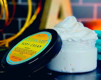 May include: A black jar with a white lid filled with a white body cream. The jar has a yellow and blue label that reads "Ocean Mist Body Cream" and lists the ingredients.