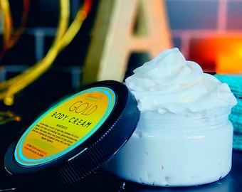 May include: A jar of white body cream with a black lid. The lid has a gold label with the words "Gold Body Cream" printed on it. The jar is sitting on a dark surface.