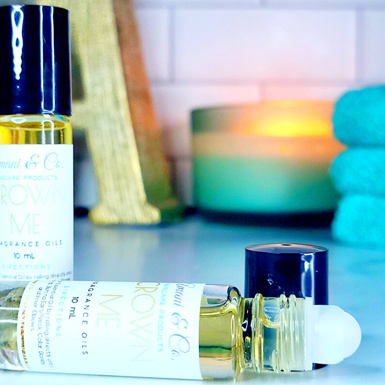 May include: Two 10 ml roll-on bottles of fragrance oil labeled "Crown Me" with black caps and gold lettering. The bottles are on a wooden surface with a candle and towels in the background.