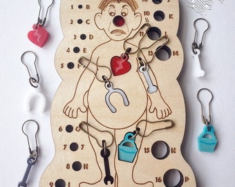 May include: A light-colored wooden needle gauge board shaped like a human figure with engraved numbers, letters, and a ruler. It includes several metal stitch markers with attached charms in shapes like hearts, bones, buckets, and wrenches.