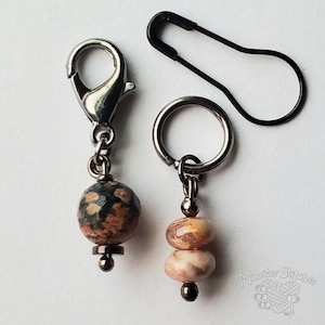 May include: Two stitch markers with metal findings and stone beads. One features a silver-tone lobster clasp with a round, mottled black and orange bead. The other has a silver-tone ring with two small, smooth, pink and tan beads. A black safety pin is included.