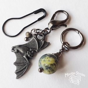 May include: A set of stitch markers including a black safety pin, a dark metal lobster clasp charm featuring a textured bat with small metallic beads, and a dark metal ring charm with a round, mottled yellow and grey stone bead. Monster Stitches.