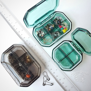 May include: Two clear plastic containers with multiple compartments, each filled with small colorful beads and jewelry findings. The containers are open and sitting on a white surface next to a ruler.