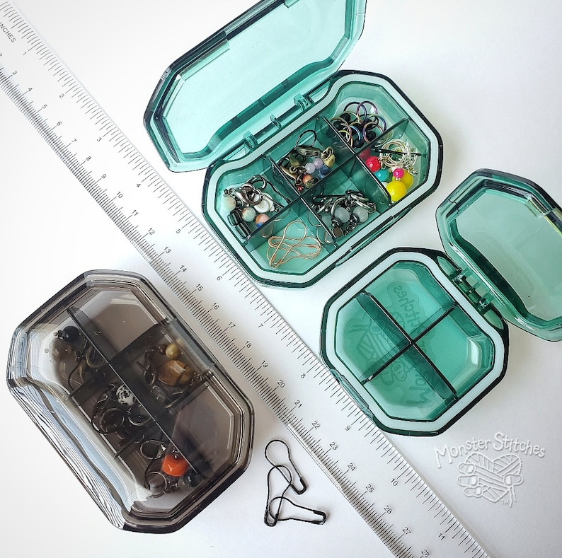 May include: Two clear plastic containers with multiple compartments, each filled with small colorful beads and jewelry findings. The containers are open and sitting on a white surface next to a ruler.