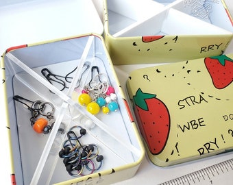 May include: A yellow and white tin box with a strawberry pattern and the words "STRAY" and "WBE" on the side. The box is open and contains a variety of colorful beads, rings, and safety pins.
