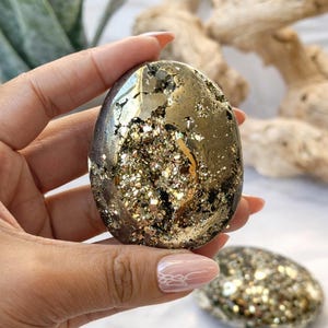 May include: A polished pyrite stone, oval-shaped, with a metallic gold and silver luster. The stone has a cavity filled with sparkling crystals. Additional pyrite stones are visible in the background, suggesting a collection of similar items.