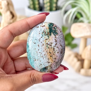 May include: An oval-shaped polished stone featuring a mottled pattern of teal, forest green, tan, and lavender hues. The surface is covered in small, dark brown circular spots and speckles, creating a natural, multi-coloured appearance.