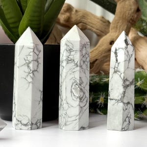 May include: Three white marble obelisks with gray veining, each with a pointed top. The obelisks are displayed on a white surface, with green plants and a piece of driftwood in the background. The obelisks are a decorative item.