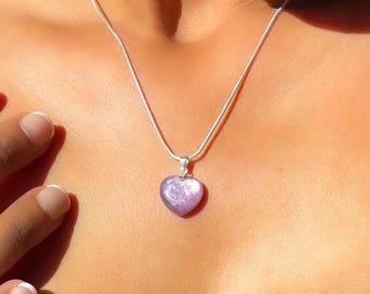 May include: A silver necklace with a heart-shaped pendant. The pendant is a light purple color and appears to be made of a translucent material. The necklace is worn around the neck.
