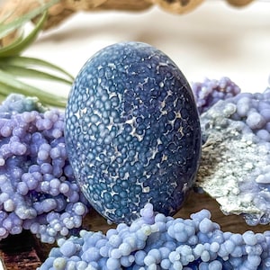 May include: A collection of blue and purple geodes and crystals. A large, oval-shaped, dark blue geode with a textured surface is the focal point. Surrounding it are clusters of light purple, grape-like formations, all displayed on a wooden tray.