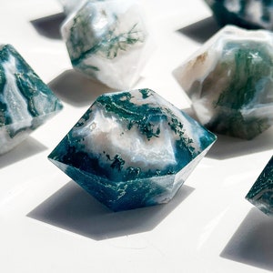 May include: Several faceted gemstone dice, each displaying a unique pattern of white, teal, and green, resembling moss. The dice are arranged on a white surface, with shadows cast by the sunlight, creating a striking display.