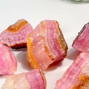 May include: A collection of raw pink cobalto calcite stones with varying shades of pink, orange, and brown. The stones have a layered, banded appearance and are displayed on a white surface. The natural crystals are a light pink color.