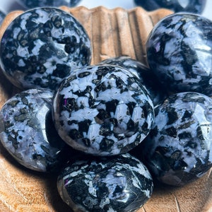 May include: A collection of tumbled snowflake obsidian stones in a wooden bowl. The stones are black with white flecks and have a smooth, polished finish.