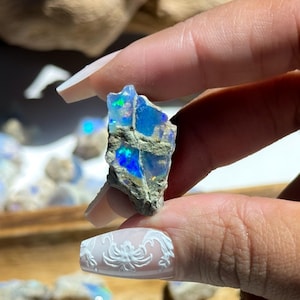 May include: A rough-cut opal gemstone with vibrant blue, green, and iridescent flashes. The stone is held between fingers, showcasing its natural, unpolished texture. Other opals are visible in the background, suggesting a collection of raw gemstones.