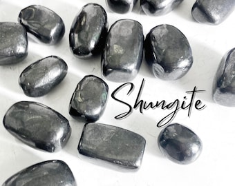 May include: A collection of smooth, black Shungite stones. The stones are various sizes and shapes, and they are arranged on a white surface.