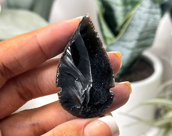 Puede incluir: Una punta de flecha de obsidiana negra con una superficie lisa y pulida. La punta de flecha se sostiene en una mano, mostrando su forma única y su color oscuro.