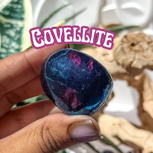 May include: A polished, oval-shaped covellite gemstone. The stone has a deep blue hue with streaks of purple. The word "COVELLITE" is displayed in a pink and white font above the stone.
