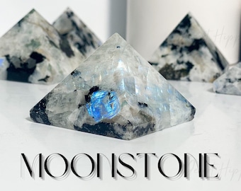 Puede incluir: Varias pirámides de piedra lunar con apariencia translúcida y cristalina. Las pirámides tienen una mezcla de tonos negros, blancos y azules. La palabra "MOONSTONE" está impresa en una fuente negra y estilizada.