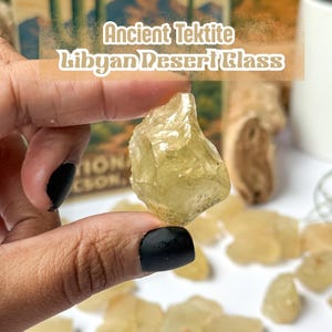 Puede incluir: Un trozo de vidrio del desierto libio de tectita antigua, sostenido entre los dedos, es el punto focal. La piedra translúcida, de color amarillo verdoso, está rodeada de otras piezas similares. El fondo incluye una ilustración de paisaje desértico y el texto "Ancient Tektite Libyan Desert Glass."