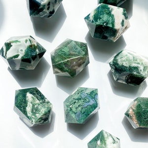 May include: A selection of hexagonal-shaped moss agate crystals. The crystals feature a mix of translucent white and opaque green patterns. The crystals are arranged on a white surface, with shadows adding depth to the composition.