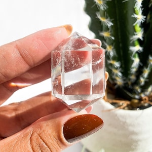May include: A clear quartz crystal point with a faceted, geometric design. The crystal is held in a hand, with a cactus in the background. The crystal is approximately 2 inches in length.