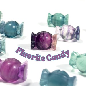 Puede incluir: Varias esculturas de fluorita con forma de caramelos en tonos morados, azules y verdes. Se muestra el texto "Fluorite Candy". Los caramelos están dispuestos sobre un fondo blanco.