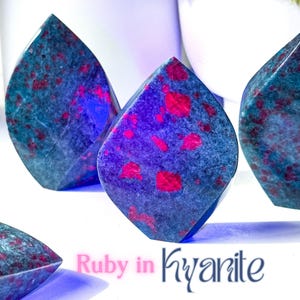 Könnte beinhalten: Vier polierte Kyanit-Steine mit Rubin-Einschlüssen. Die Steine sind tiefblau mit roten Flecken durchzogen. Die Steine sind tropfenförmig und in einer Reihe auf einer weißen Oberfläche angeordnet. Der Text "Ruby in Kyanite" ist in rosa Buchstaben unter den Steinen geschrieben.
