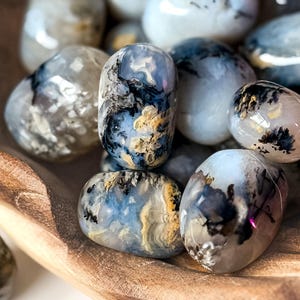 May include: A collection of polished, tumbled stones featuring a translucent white base with intricate, branching black dendrite inclusions and patches of soft yellow and blue mineral deposits, all gathered together in a smooth wooden bowl.