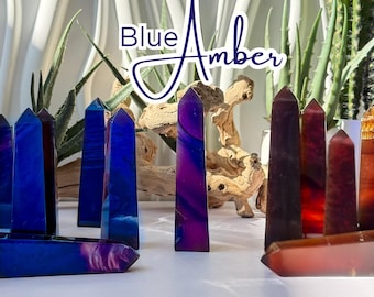 May include: A collection of polished crystal obelisks in shades of blue, purple, and red. The crystals are arranged on a white surface, with the text "Blue Amber" in the background. The obelisks vary in size and shape, with a natural wood element and plants in the background.