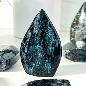May include: A polished, teardrop-shaped stone carving featuring a deep black base colour with prominent, shimmering blue, fibrous inclusions that create a reflective, metallic-like pattern across the surface of the dark, smooth mineral specimen.