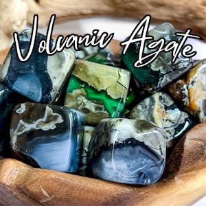 May include: A collection of polished Volcanic Agate stones in shades of green, blue, and brown, displayed in a wooden bowl. The stones have unique patterns and textures, with the words "Volcanic Agate" in a white script.
