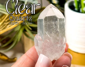 Könnte beinhalten: Klare Quarzkristallspitze mit natürlicher, trüber Optik. Der Kristall wird in einer Hand gehalten und der Text "Klarer Quarz Klarheit + Fokus" ist im Hintergrund sichtbar.