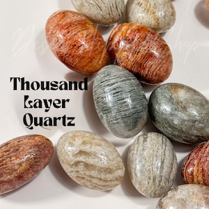 May include: A collection of polished, oval-shaped thousand layer quartz stones in shades of gray, brown, and red. The stones have a layered, striated pattern.
